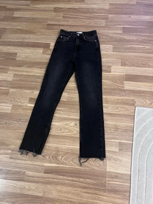 Svarta jeans med slits - Säljer ett par svarta jeans med hög midja och raka ben. Jeansen har trendiga råa, fransiga kanter nertill och slitsar på sidorna vid bensluten. Klassisk femficksmodell och stängs med knapp och dragkedja. Perfekta för en cool och avslappnad stil.