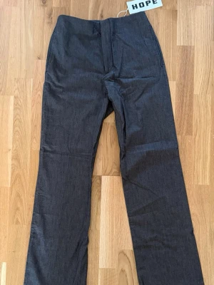 HOPE Mörkgrå kostymbyxor med rak passform - Snygga mörkgrå linnebyxor med rak passform och klassisk look. Byxorna har en bakficka med knapp och diskreta sidfickor framtill. Materialet är svalt och lätt, perfekt för en stilren outfit. Inget synligt märke på byxorna.