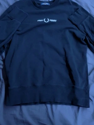 Svart sweatshirt från Fred Perry - En svart sweatshirt från Fred Perry med vit broderad logotyp på bröstet. Tröjan har rund halsringning och ribbade muddar vid ärmslut och nederkant. Materialet är mjukt och bekvämt, perfekt för en avslappnad stil. seriöst bara använd en gång och är i perfekt skick