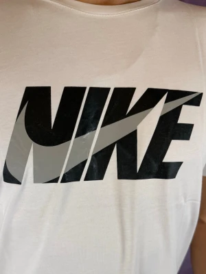 Vit Nike t-shirt med stor logga - Snygg vit t-shirt från Nike med stor svart och grå logga på bröstet. Klassisk rund halsringning och korta ärmar. Tillverkad i mjuk bomull som känns skön mot huden. Perfekt för en sportig och avslappnad stil.