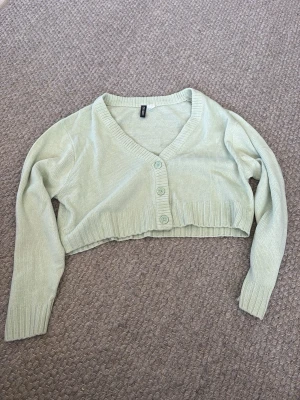 Ljusgrön cropped kofta från H&M - Säljer en ljusgrön cropped kofta från H&M med v-ringning och tre knappar framtill. Koftan har långa ärmar och ribbade muddar vid ärmslut och nederkant. Perfekt att slänga över en topp för en chill look.