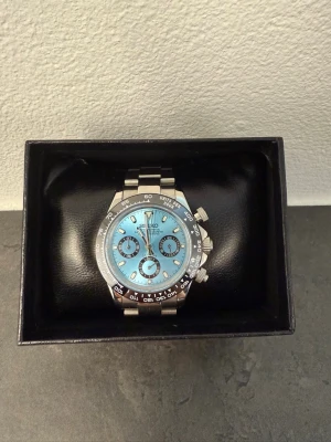 Seiko silverklocka med blå urtavla - Säljer en snygg Seiko klocka med silverfärgat armband och boett. Urtavlan är ljusblå med tre mindre svarta cirklar och tydliga index. Klockan har kronograf-funktion och sportig design, perfekt för dig som gillar stilrena accessoarer.