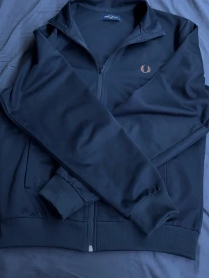 Svart zip-tröja från Fred Perry - Cool svart zip-tröja från Fred Perry med klassisk laurel-logga broderad på bröstet. Tröjan har hög krage, ribbade muddar och dragkedja framtill. Perfekt för en sportig och avslappnad look. Materialet är mjukt och stretchigt. använd ett par tre gånger men är i nyskick fortfarande utan skador