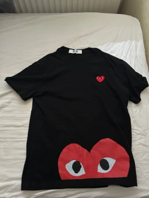 Svart Comme des Garçons Play t-shirt - Svart t-shirt från Comme des Garçons Play med ikoniskt rött hjärta med ögon på bröstet och ett stort hjärta med ögon nertill. Klassisk passform och rund hals. Perfekt för dig som gillar streetwear och vill sticka ut med en cool detalj.