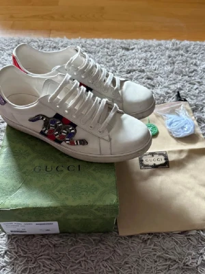 Gucci Ace  - Säljer ett par vita Gucci Ace sneakers med ikonisk orm-applikation i pärlor och paljetter på sidan. Skorna har klassisk låg profil, vita snören och detaljer i rött och blått på hälen. Kommer med originalkartong och dustbag. Liten defekt tån på skon där sömmarna lossnar. Inget man märker 