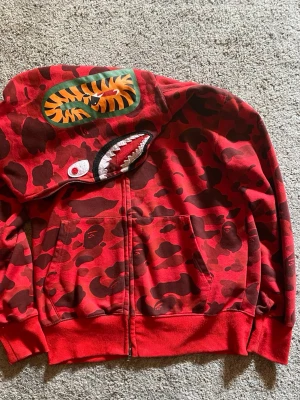Bape hoodie röd - Nyskick då den är använd väldigt sparsamt, möjligtvis något katthår på den men tar bort det innan utskick.       Det står att storleken är L men sitter som en M.               Vid frågor om hoodien skriv gärna privat💯