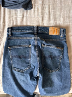 Blå jeans från Nudie Jeans - Säljer ett par klassiska blå jeans från Nudie Jeans med fem fickor och snygga kontrastsömmar. Jeansen har normal midja och raka ben, tillverkade i slitstark denim. Perfekta för en avslappnad och trendig look. Modell: grim tim/ splitternya!