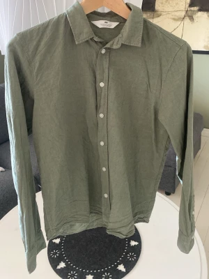 Grön linneskjorta från H&M - Snygg grön skjorta från H&M i linne med klassisk krage och knäppning framtill. Skjortan har lång ärm och en avslappnad passform, perfekt för en stilren look. Enkel design utan mönster eller extra detaljer.