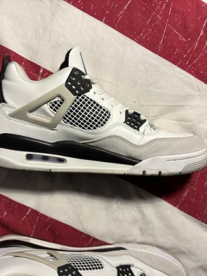 Vita Air Jordan 4 sneakers - Snygga Air Jordan 4 sneakers i vitt med svarta och grå detaljer. Skorna har meshpaneler på sidorna, ikoniska plastvingar och synlig Air-enhet i sulan. Ovandelen är i läder och mocka, med klassisk siluett och cool design som sticker ut.