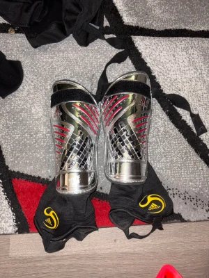 Adidas benskydd silver och svart - Säljer ett par Adidas benskydd i silver med svarta och röda detaljer. De har justerbara svarta kardborreband och medföljande svarta strumpor med gul Adidas-logga. Perfekta för fotbollsträning eller match.