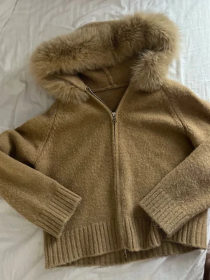 Beige jacka med pälshuva - Mysig beige jacka med dragkedja och stor huva kantad med fluffig fuskpäls. Jackan har ribbade muddar vid ärmslut och nederkant, samt en mjuk och lurvig yta som ger en trendig look. Perfekt för kyliga dagar när du vill vara både varm och snygg.
