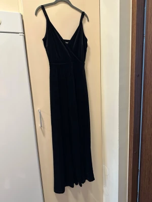 Svart sammetsjumpsuit med smala axelband från bikbok - Stilren svart jumpsuit i mjuk sammet med smala axelband och omlott-ringning. Vida ben och markerad midja ger en snygg siluett. Perfekt för dig som vill ha en trendig och bekväm look.
