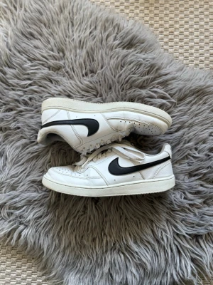 Vita Nike sneakers med svart swoosh - Säljer ett par klassiska vita Nike sneakers med svart swoosh på sidan. Skorna har lågt skaft, vita snören och ovandel i läder. Perfekta för dig som gillar en clean och tidlös stil.