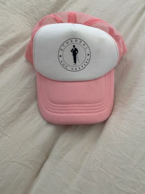 Rosa och vit keps från Ethereal - Snygg rosa och vit keps från Ethereal med mesh-bakstycke och tryckt logga framtill. Framsidan är vit med svart tryck och baksidan är i rosa mesh för extra ventilation. Perfekt accessoar för en cool och avslappnad stil.