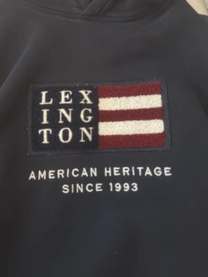 Mörkblå hoodie från Lexington - Snygg mörkblå hoodie från Lexington med broderad logga och amerikanskinspirerad flagga på bröstet. Klassisk huva och mjukt material som känns skönt mot huden. Perfekt för dig som gillar stilren och tidlös design.