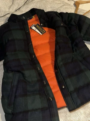 Rutig pufferjacka från Polo Ralph Lauren - Säljer en riktigt snygg pufferjacka från Polo Ralph Lauren i mörkgrönt och svart rutigt tyg med orange foder. Jackan har tryckknappar, klassisk krage och två sidofickor. Perfekt för kalla vinterdagar och ger en stilren look.