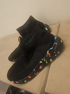 Svarta sneakers från Wozniak - Säljer ett par svarta sneakers från Wozniak med stickad ovandel och högt skaft. Sulan är chunky och har färgglada detaljer i blått, orange och gult. Perfekt för dig som gillar en unik och modern look.