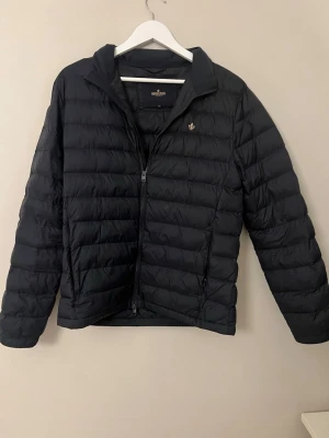 Svart dunjacka från Morris Stockholm - Säljer en svart dunjacka från Morris Stockholm med quiltad design och guldfärgad logotyp på bröstet. Jackan har dragkedja framtill och stårkrage. Perfekt för kalla vinterdagar och snygg att matcha med jeans eller sneakers.
