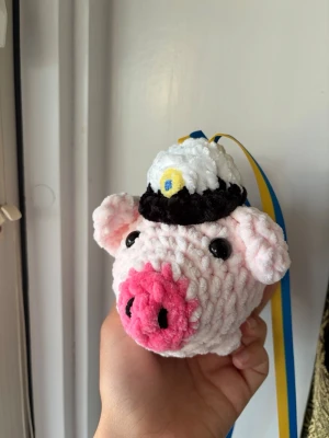 Studentgris i mjukt garn - En handgjord rosa gris i mjukt garn med en vit och svart studentmössa på huvudet. Grisen har stora svarta ögon, rosa nos och öron, samt ett blågult band fäst i mössan. Perfekt som studentpresent eller dekoration.