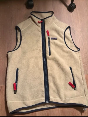  Fleeceväst från Patagonia - Snygg fleeceväst från Patagonia med marinblå detaljer och röda dragkedjor. Västen har hög krage, tre fickor med dragkedja och är tillverkad i mjukt fleece-material. Perfekt lager-på-lager-plagg för kyliga dagar. Använd ett fåtal gånger, väldigt bra skick!