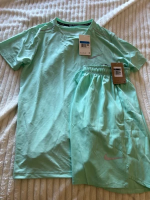 Nike Miller set - Snyggt mintgrönt träningsset från Nike med t-shirt och shorts. Båda plaggen har diskret Nike-logga i silver och är tillverkade i ett lätt, snabbtorkande material som passar perfekt för löpning eller gym. Setet har en sportig och fräsch look.
