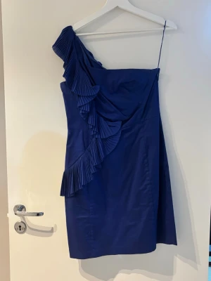 Blå one shoulder klänning med volang - Stilren blå kortklänning med one shoulder och plisserad volangdetalj som går från axeln ner över framsidan. Klänningen har smal axelrem och ärmlös design, vilket ger en modern och trendig look. Tillverkad i ett slätt material som ger en snygg siluett.