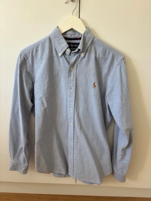 Blå skjorta från Ralph Lauren - Klassisk ljusblå skjorta från Ralph Lauren med button-down krage och broderad logga på bröstet. Skjortan har lång ärm, knappar framtill och är i en skön bomullskvalitet. Perfekt för en stilren look.