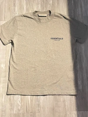 Essentials Fear of God Tee - Säljer en ljusgrå Essentials t-shirt i nyskick med tagsen kvar. Tillverkad i kraftig bomull med en snygg oversized passform. Klassisk gummilogga på bröstet och patch i nacken. 