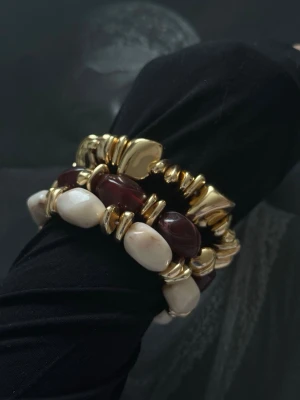 Chunky armband med pärlor och guldton - Säljer ett chunky armband med stora pärlor i vinrött, vitt och guldfärgade detaljer. Armbandet har en cool och unik design med oregelbundna former och elastisk tråd som gör det lätt att ta på. Perfekt för dig som vill sticka ut med accessoarer.