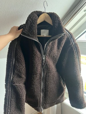 Mörkbrun populär teddyjacka från Hm - Mysig mörkbrun teddyjacka med hög krage och hel dragkedja framtill. Jackan har två sidofickor och detaljer i brunt skinnimitation längs ärmarna. Använd fåtal gånger. Slutsåld