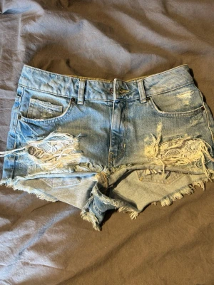 Blå slitna jeansshorts med fransar - Säljer ett par ljusblå jeansshorts med slitna detaljer och fransig kant. I hålen är det ett fint vitt spetts mönster som täcker.