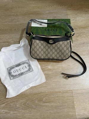  Supreme axelremsväska - Snygg handväska i beige canvas med klassiskt GG Supreme-mönster och svarta läderdetaljer. Väskan har guldfärgad GG-logga, dragkedja upptill och justerbar axelrem. Perfekt för dig som vill ha en ikonisk och trendig accessoar.