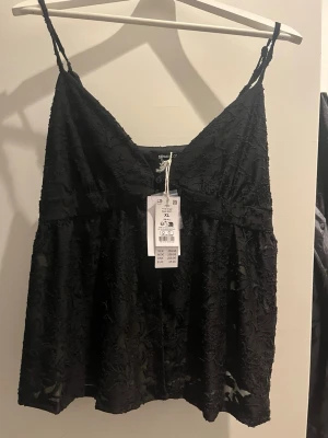 Svart spetslinnetopp från Gina Tricot NY - Svart linnetopp från Gina Tricot med tunna axelband och v-ringning. Toppen har ett vackert spetsmaterial med blommiga detaljer och är lätt utsvängd nertill. Perfekt för att skapa en trendig och stilren look. NY MED PRISLAPP