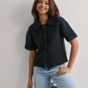 Marinblå stickad skjorta Gina Tricot - Stickad skjorta i mörkgrått från Gina Tricot med klassisk krage och korta ärmar. Skjortan har fyra knappar framtill och är gjord i ett mjukt, stickat material som ger en chill och cozy vibe. Perfekt för dig som gillar enkel och stilren design.