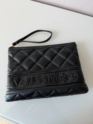 Svart kuvertväska från Valentino - Snygg svart kuvertväska från Valentino med quiltat mönster och tydlig logga framtill. Väskan har en avtagbar handledsrem och är tillverkad i konstläder med lyxig känsla. Perfekt för att bära det viktigaste med stil.