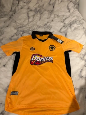 Wolves retro fotbollströja Doritos - Gul och svart retro fotbollströja från Wolves med Doritos-logga på bröstet. Tröjan har korta ärmar, svart krage och paneler på sidorna. Tillverkad i lätt och ventilerande polyester, perfekt för träning eller match.