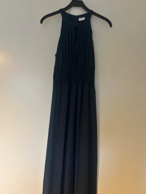 Mörkblå långklänning från VILA - Elegant mörkblå långklänning från VILA med hög halsringning och spets i midjan. Klänningen har ett mjukt, flytande tyg och är ärmlös med en stilren siluett. Perfekt för speciella tillfällen där du vill känna dig extra fin som tex som balklänning. Använd endast vid två tillfällen säljes på grund av att den inte används.