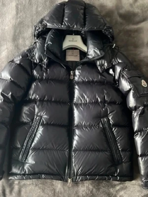 Svart Moncler Maya  - Snygg svart dunjacka från Moncler med glansigt material och klassisk logotyp på ärmen. Jackan har huva, två dragkedjeförsedda fickor och quiltad design. Perfekt för kalla vinterdagar och riktigt trendig stil. Funkar till M
