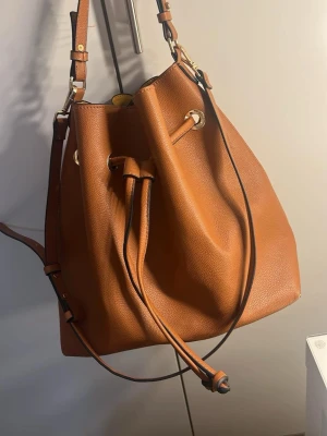 Brun bucket bag med dragsko - Snygg brun axelväska i bucket-modell med justerbar axelrem och dragskostängning. Väskan har guldfärgade metalldetaljer och är gjord i ett slätt, läderliknande material. Perfekt för dig som gillar stilrena och trendiga accessoarer.