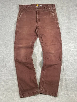 Carhartt Workwear Straight Fit Pants Faded Brick Brown W32 - Carhartt straight fit workwear pants in a faded brick brown tone. Clean worn-in look with natural fading and solid structure. Easy everyday pair with a mid-rise and straight leg, works with both sneakers and boots.  Waist 42 cm, length 75.5 cm, rise 27 cm, thigh 28 cm, knee 21 cm, leg opening 18 cm.  Take your favorite pants and measure them the same way as shown in the photos to ensure a good fit.  Used condition with visible wear, see photos.