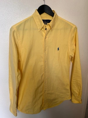 Gul skjorta från Ralph Lauren - Klassisk gul skjorta från Ralph Lauren med blå broderad logga på bröstet. Slim fit-modell med knappar hela vägen och lång ärm. Perfekt för dig som gillar stilrena och färgglada plagg.