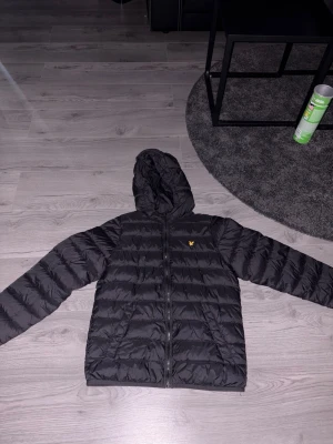 Svart pufferjacka från Lyle & Scott - Svart pufferjacka med huva från Lyle & Scott. Jackan har dragkedja framtill, två sidofickor och det klassiska gula logomärket på bröstet. Quiltad design som håller värmen och ger en snygg siluett. Perfekt för kalla dagar.
