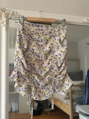 Blommig kortkjol med volang och snörning - Supersöt kortkjol med blommigt mönster i lila, gul och grön på vit botten. Kjolen har volangkant och snörning framtill för en trendig look. Perfekt för vår och sommar, lätt och luftig i materialet.