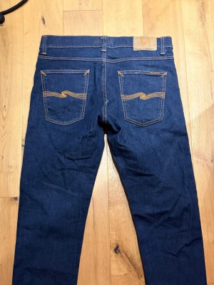 Mörkblå jeans från Nudie  - Säljer ett par mörkblå jeans från Nudie Jeans med klassisk femficksdesign och snygga orangea sömmar. Jeansen har raka ben och är tillverkade i kraftigt denimtyg. Perfekta för dig som gillar stilrena och tidlösa jeans. Står ingen storlek vad jag kan se men mäts till c.a W30 & L31/32. Nästintill nytt skick men har två artificiella slitningar.