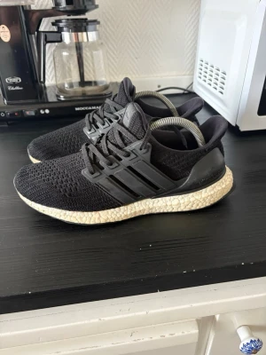 Adidas Ultra Boost svarta sneakers - Svarta Adidas Ultra Boost sneakers med stickad ovandel och klassiska tre ränder på sidan. Skorna har en vit Boost-sula som ger grym komfort och respons. Ovandelen är i mesh för bra andningsförmåga och passform. Perfekta för dig som gillar sportig och stilren look.