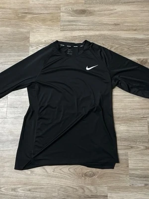 Svart långärmad träningströja Nike - Svart långärmad träningströja från Nike med vit logga på bröstet. Tillverkad i ett lätt och stretchigt funktionsmaterial som andas, perfekt för träning och sportaktiviteter. Enkel och stilren design med rund hals.