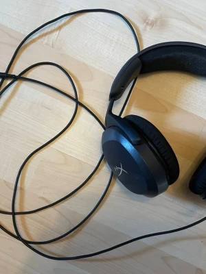 Svarta HyperX over-ear hörlurar - Säljer ett par svarta over-ear hörlurar från HyperX med kabelanslutning. Hörlurarna har mjuka öronkuddar i tyg och plast, samt en vadderad bygel för extra komfort. Perfekta för gaming eller musiklyssning.