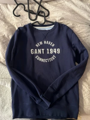 Mörkblå sweatshirt från Gant - Mörkblå sweatshirt från Gant med tryck 'New Haven Gant 1949 Connecticut' i vitt framtill. Klassisk rund halsringning och ribbade muddar vid ärmslut och nederkant. Perfekt för en avslappnad och sportig stil.
