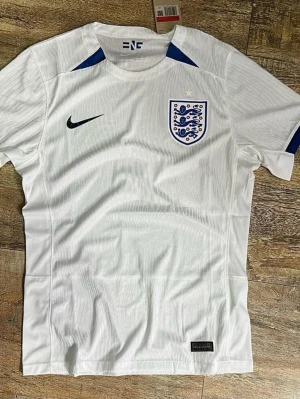 England Nike vit fotbollströja - Snygg vit fotbollströja från Nike med Englands landslagsmärke på bröstet och svart Nike-logga. Tröjan har blå detaljer vid halsen och är tillverkad i ett lätt och ventilerande material, perfekt för träning eller match.