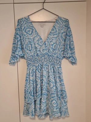 Blåvit kortklänning från Amisu  - Superfin blåvit kortklänning från Amisu med v-ringning, smockad midja och volangig kjol. Klänningen har korta ärmar och ett unikt mönster som ger en fräsch sommarkänsla. Perfekt för varma dagar och lätta outfits.
